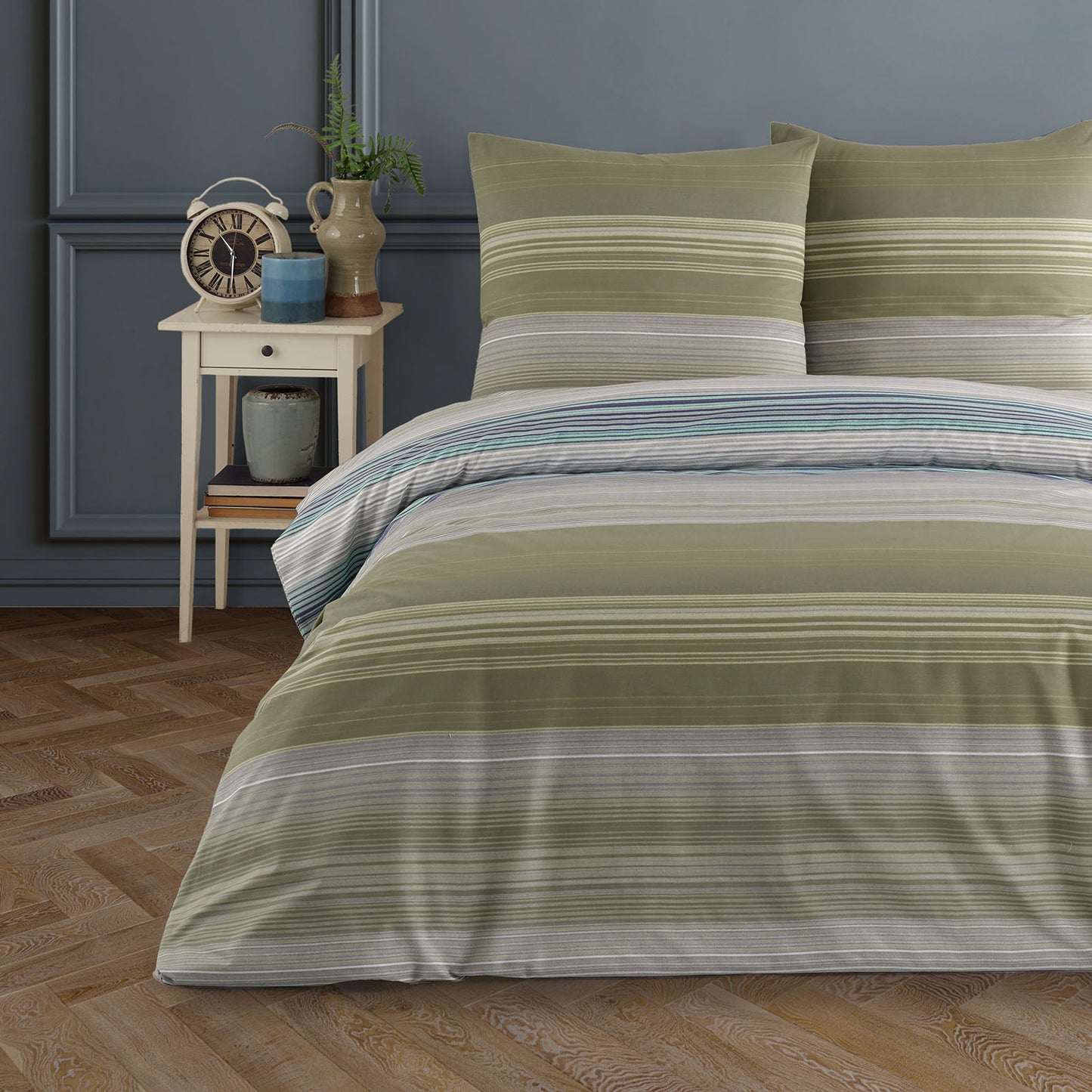 Doppelbett mit gestreifter Bettwäsche in Olivgrün, Grau und Weiß. Die Einrichtung ist klassisch mit einem Nachttisch, Vintage-Uhr, Keramikvasen und grünem Wandpaneel im Hintergrund.