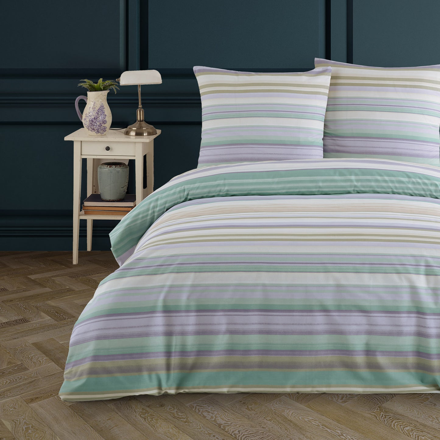 Doppelbett mit pastellfarbener Bettwäsche in feinen horizontalen Streifen in Mintgrün, Flieder, Creme und Beige. Dekoriert mit einem Nachttisch, Lampe und Lavendelkanne vor dunkelblauer Wand.